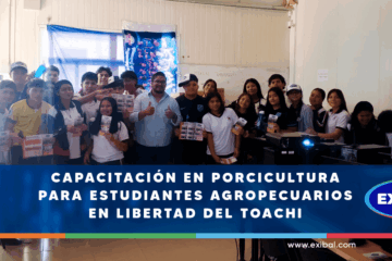 Un grupo de estudiantes agropecuarios posa junto a un instructor en un aula durante una capacitación en porcicultura organizada por EXIBAL en Libertad del Toachi. Los jóvenes sostienen folletos informativos y al fondo se observa equipo de cómputo y una pizarra.