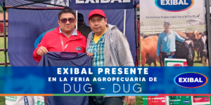 Imagen de un stand de Exibal en una feria agropecuaria, con dos personas posando frente a un fondo con banners de la marca y sosteniendo una camiseta promocional. En la parte inferior aparece una franja azul con el texto “Exibal presente en la Feria Agropecuaria de Dug-Dug”.